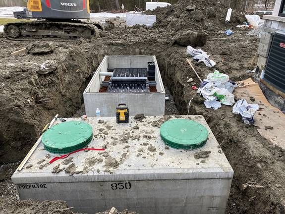 Des fosses septiques en béton sont installées sur un chantier de construction, avec une excavatrice en arrière-plan.