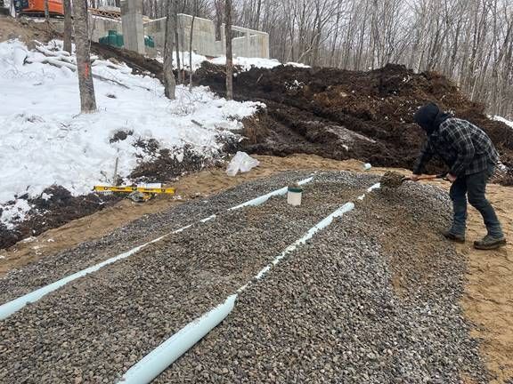 Une personne pellete du gravier sur des tuyaux de drainage dans un chantier enneigé.