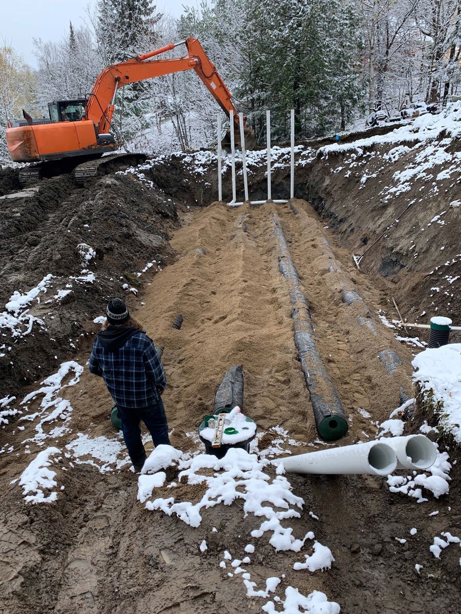 Chantier : excavatrice, homme et tuyaux dans une tranchée, probablement pour l'installation d'une fosse septique, avec un peu de neige.