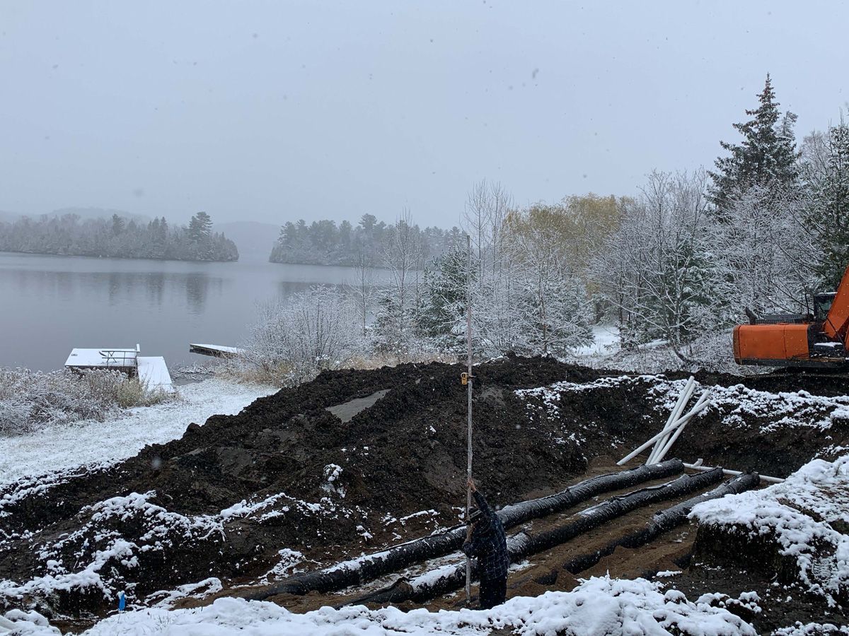 Chantier de construction sous la neige près d'un lac. Des tuyaux noirs sont dans des tranchées, une excavatrice est présente.