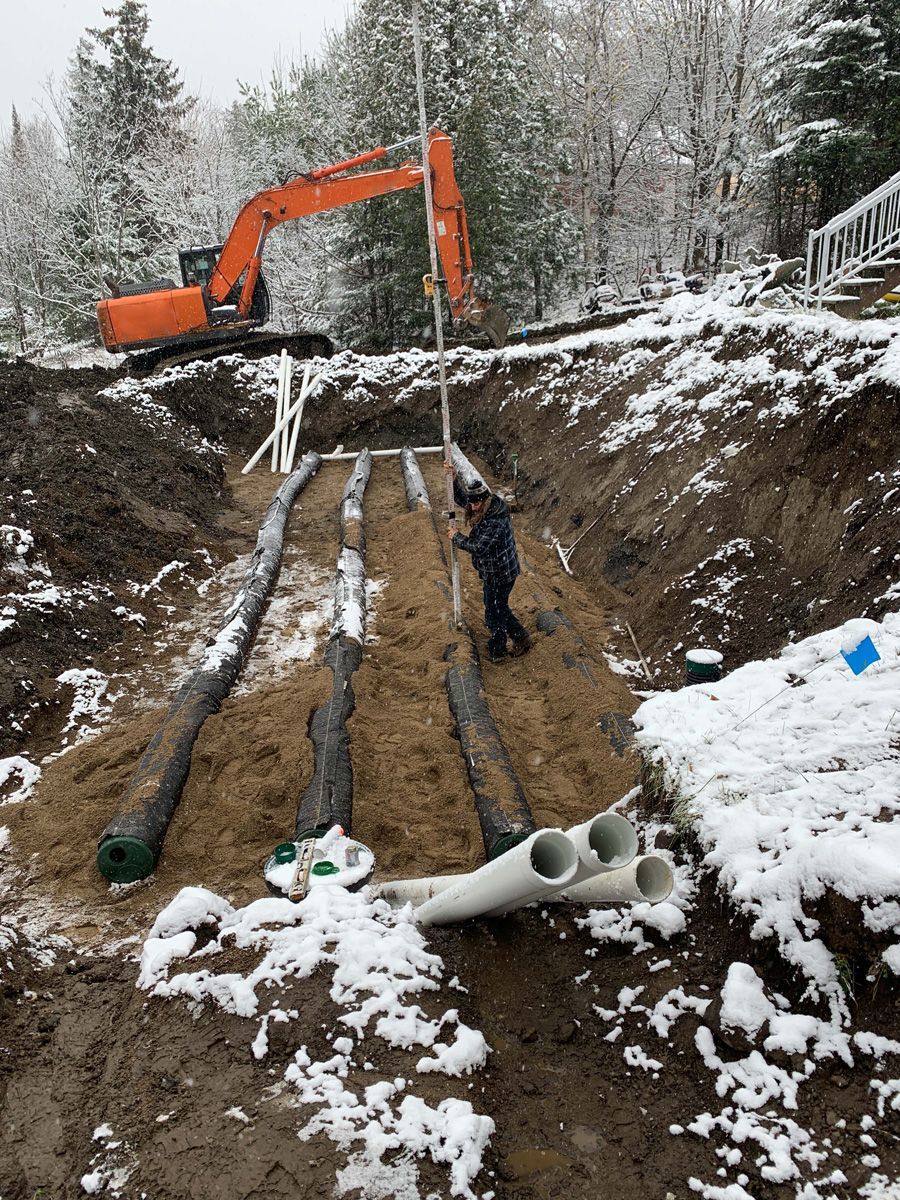 Une pelleteuse et un ouvrier installent des tuyaux de fosse septique dans un décor extérieur enneigé.
