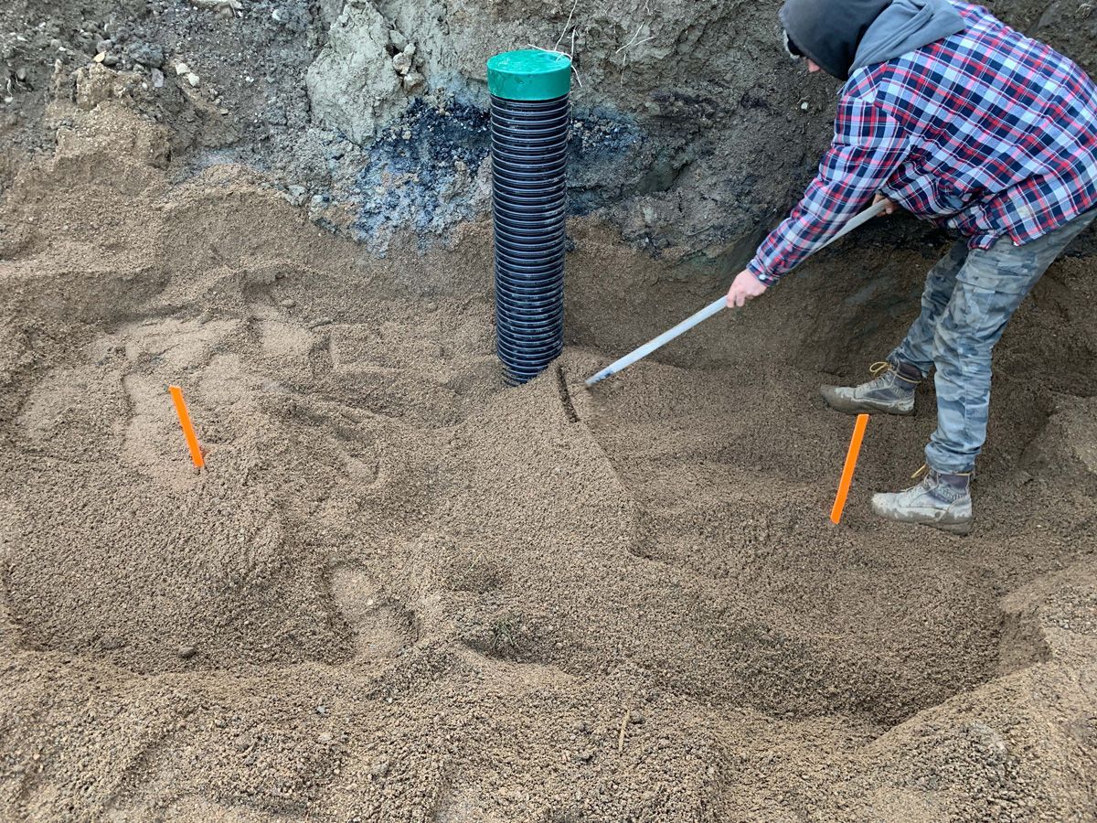 Une personne ratisse du paillis autour d'un tuyau de drainage noir. Des marques orange indiquent le périmètre.
