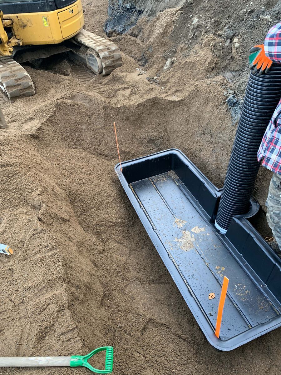 Une personne utilise un bac noir et un tuyau flexible pour tamiser la terre sur un chantier de construction près d'une petite excavatrice.