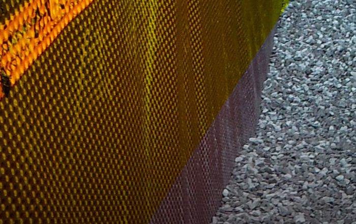 Gros plan sur une surface texturée de couleur dorée avec un motif tissé, à côté d'un lit de gravier gris.