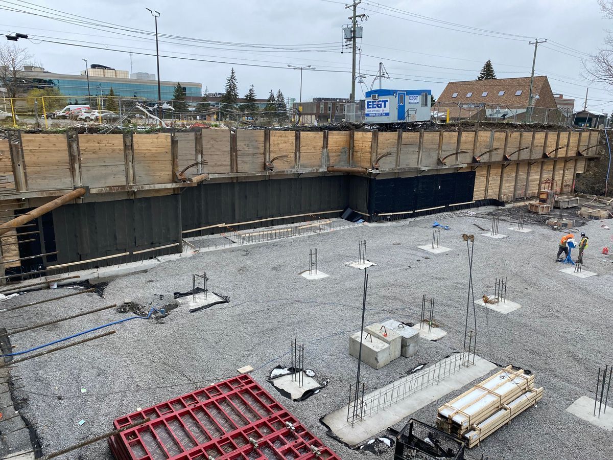 Chantier de construction avec murs de soutènement en bois, gravier, bases en béton et ouvriers.