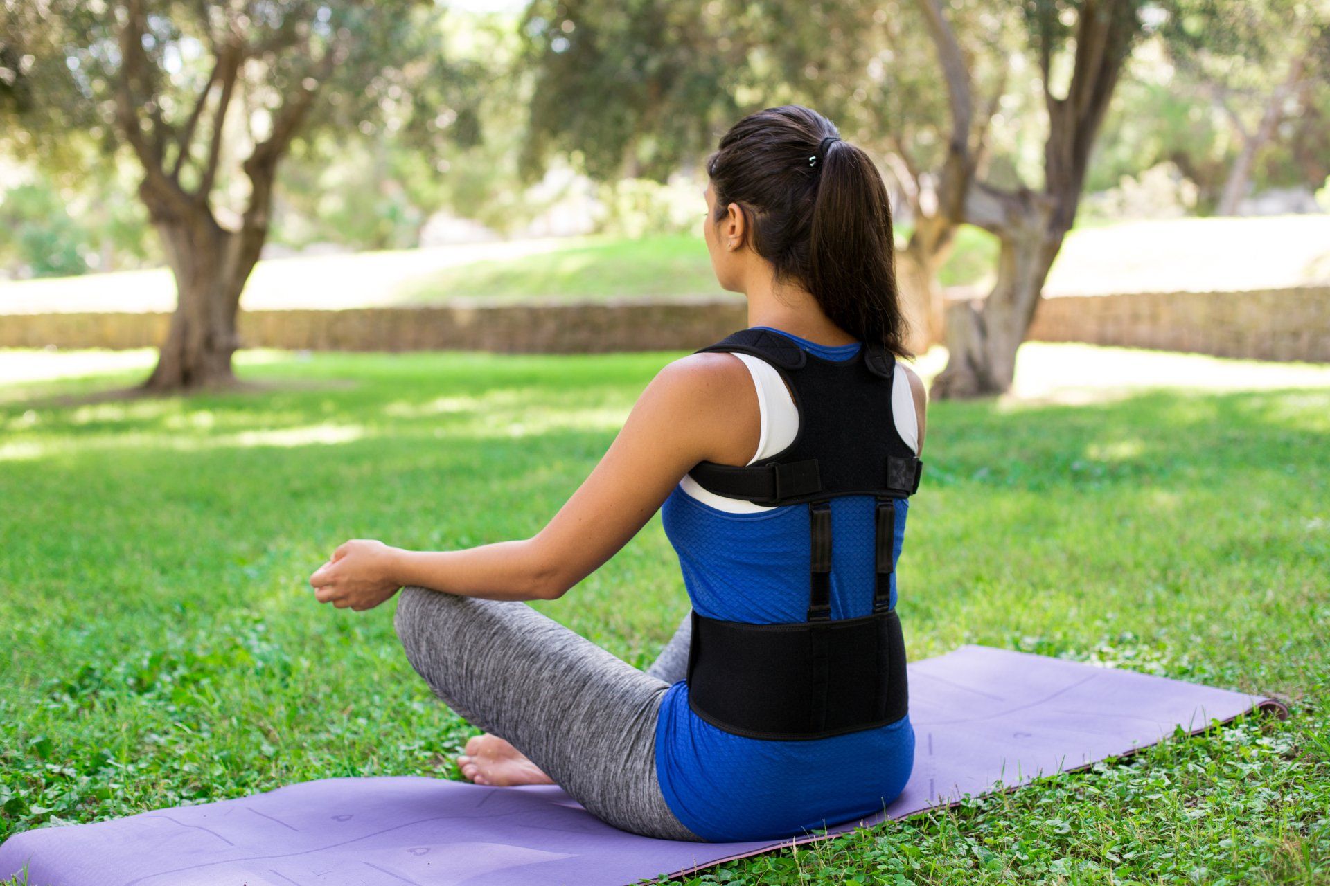 A beautiful girl in a park dressed in sportswear wearing Back posture corrector sit on yoga mat in ypoga pose.