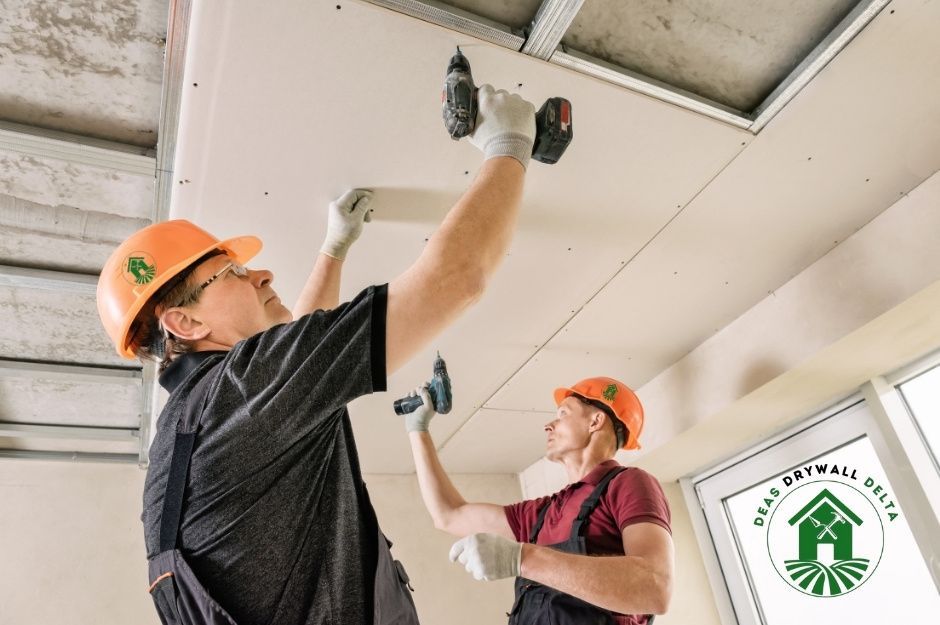 Drywall repair company workers in orange hard hats use a power drill to secure drywall panels to the ceiling while a colleague in a red hard hat assists. They're working on ceiling repairs in an unfinished residential construction site near Southborough Dr, and Biddesden Pl, BC.