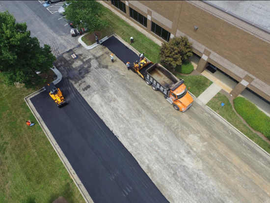 “Aerial view of workers and equipment resurfacing a paved area beside a commercial building during an asphalt service in Fulshear, TX”