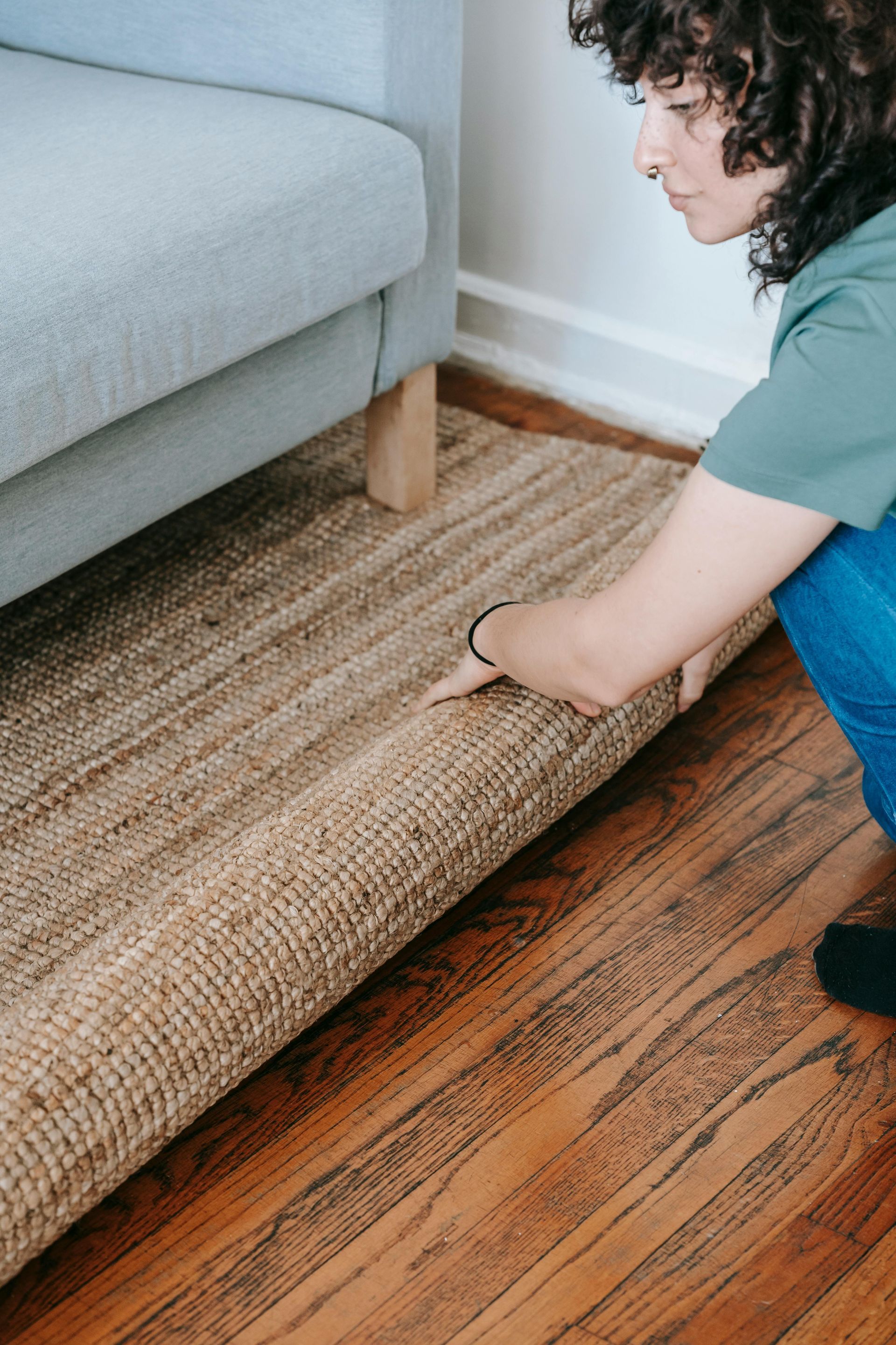A man unrolls a patterned rug on a wooden floor, while a woman in casual clothes works on a laptop on a bed. A man unrolls a patterned rug on a wooden floor, while a woman in casual clothes works on a laptop on a bed.