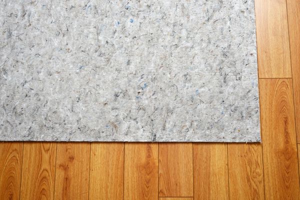 Multicolored rug is lifted to reveal a gray non-slip pad underneath on a polished wooden floor.