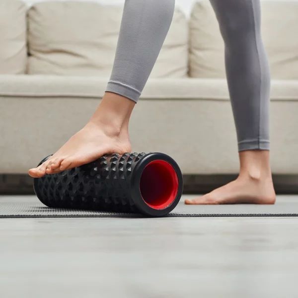 A person is standing on a yoga mat with their foot on a foam roller.