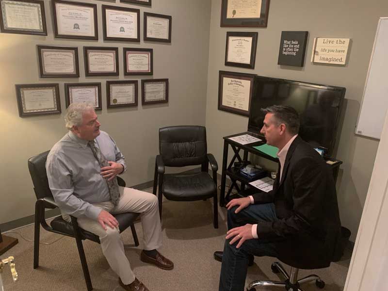 Two men are sitting in chairs in a room talking to each other.