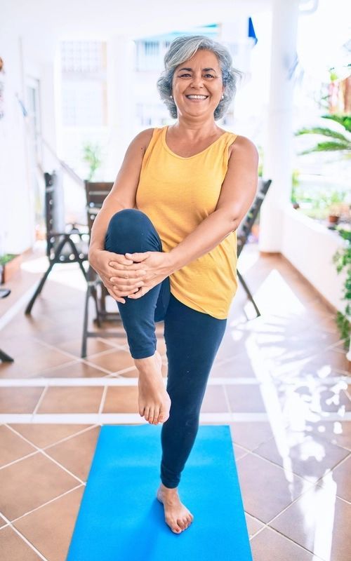 An older woman is standing on one leg on a yoga mat.