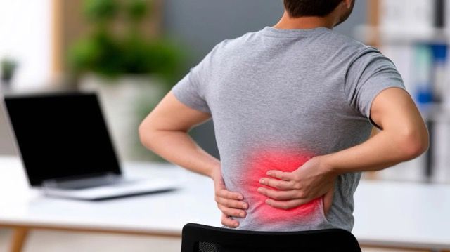 Person holding lower back in pain beside a laptop on a desk, with red pain highlight.
