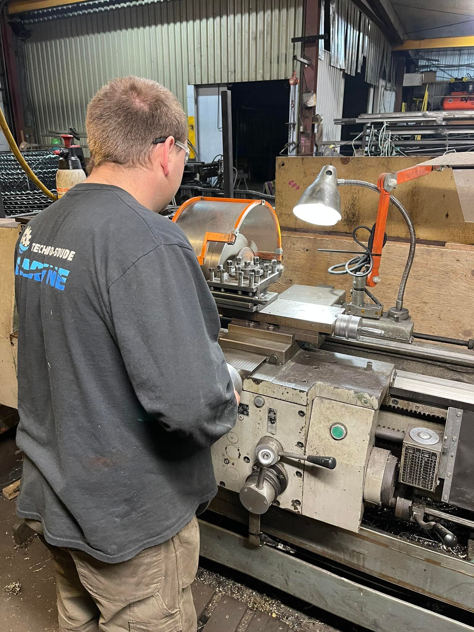 Homme travaillant sur un tour à métaux dans un atelier, vu de dos sous une lampe de chantier