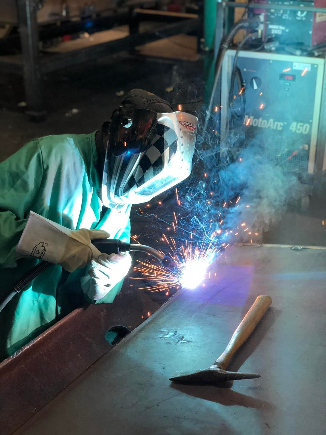 Un soudeur en chemise turquoise et visière de protection soude du métal ; des étincelles jaillissent dans l’atelier.