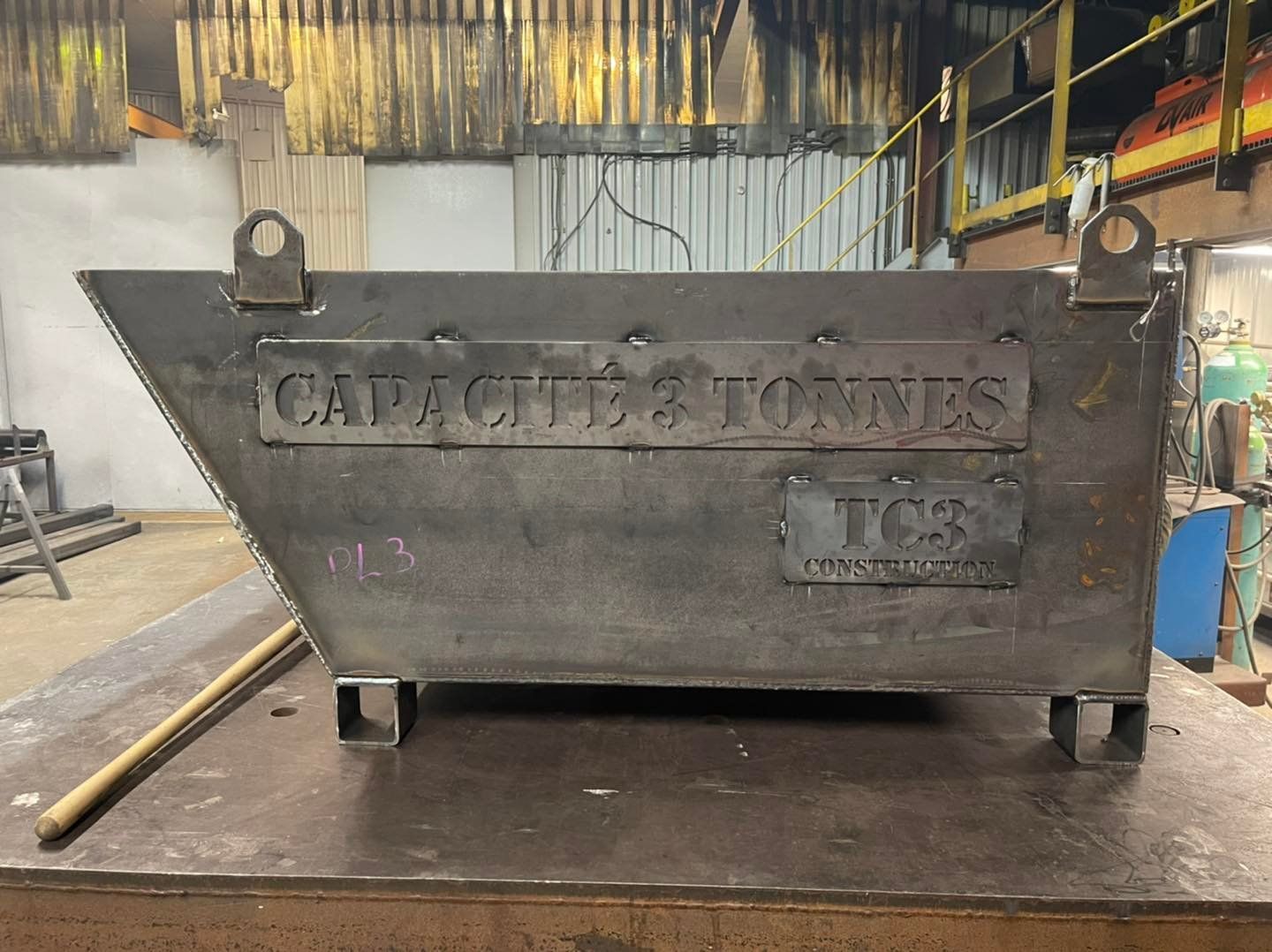 Une benne à ordures industrielle en métal rouillé, posée sur le sol d'un atelier, avec des inscriptions délavées et des poubelles bleues en arrière-plan.