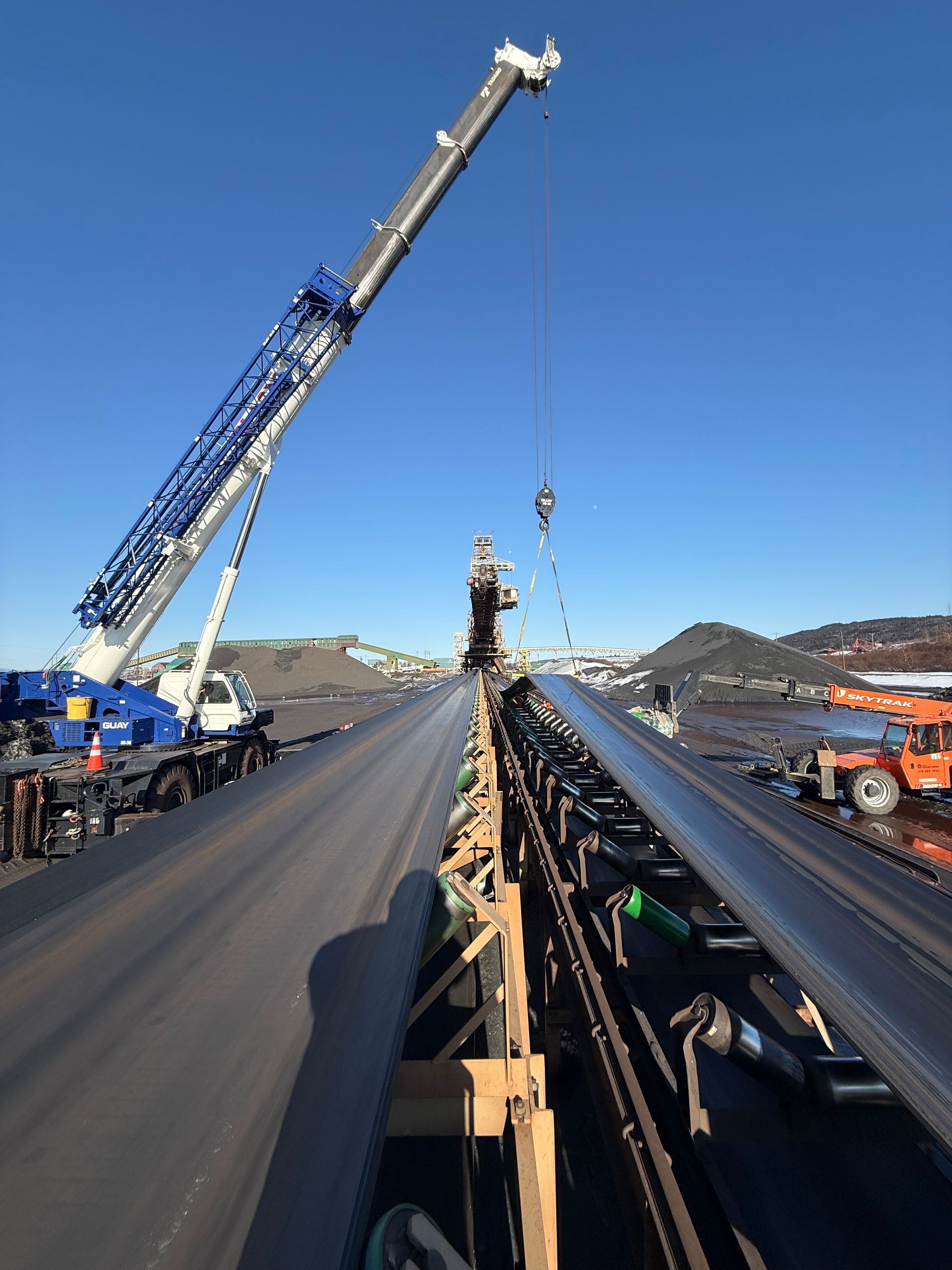 Une grue soulève des matériaux au-dessus d'un grand tuyau noir ou d'un convoyeur sur un chantier sous un ciel bleu dégagé.