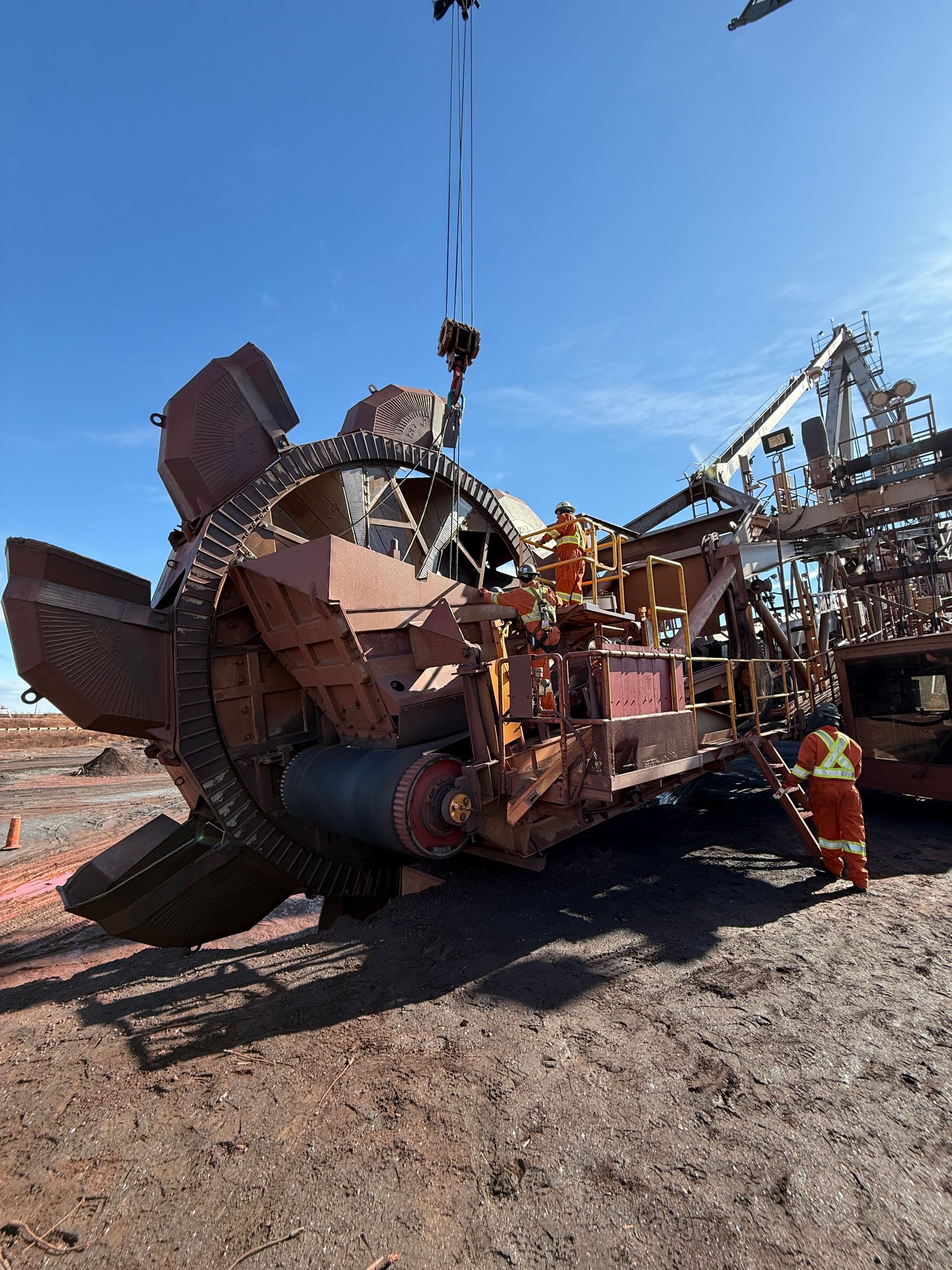Une énorme pelle mécanique broie une machine métallique rouillée sur un chantier poussiéreux, sous un ciel d'un bleu limpide.