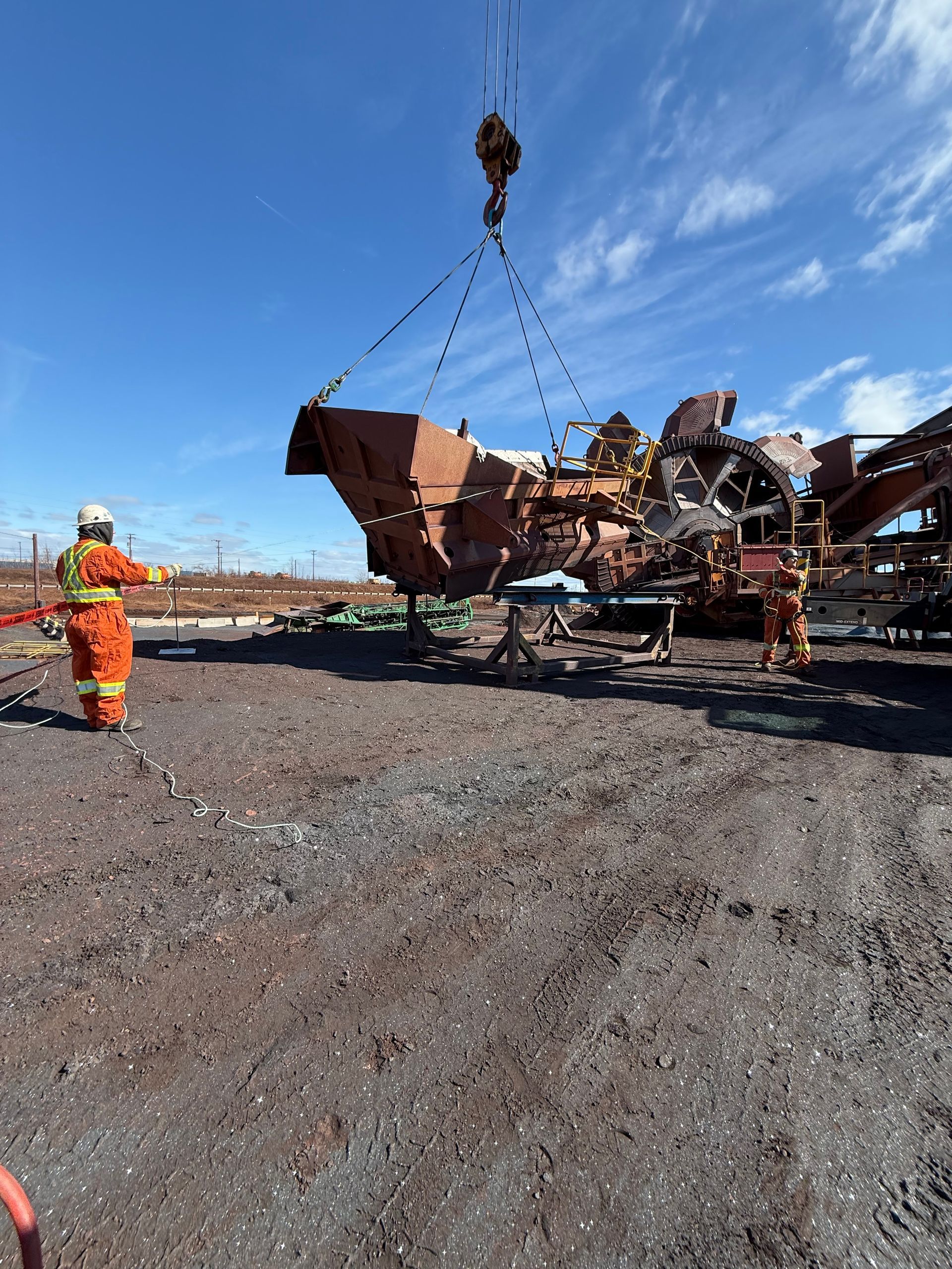 Un ouvrier en tenue orange se trouve près d'un véhicule rouillé et écrasé, soulevé par une grue sous un ciel bleu dégagé.
