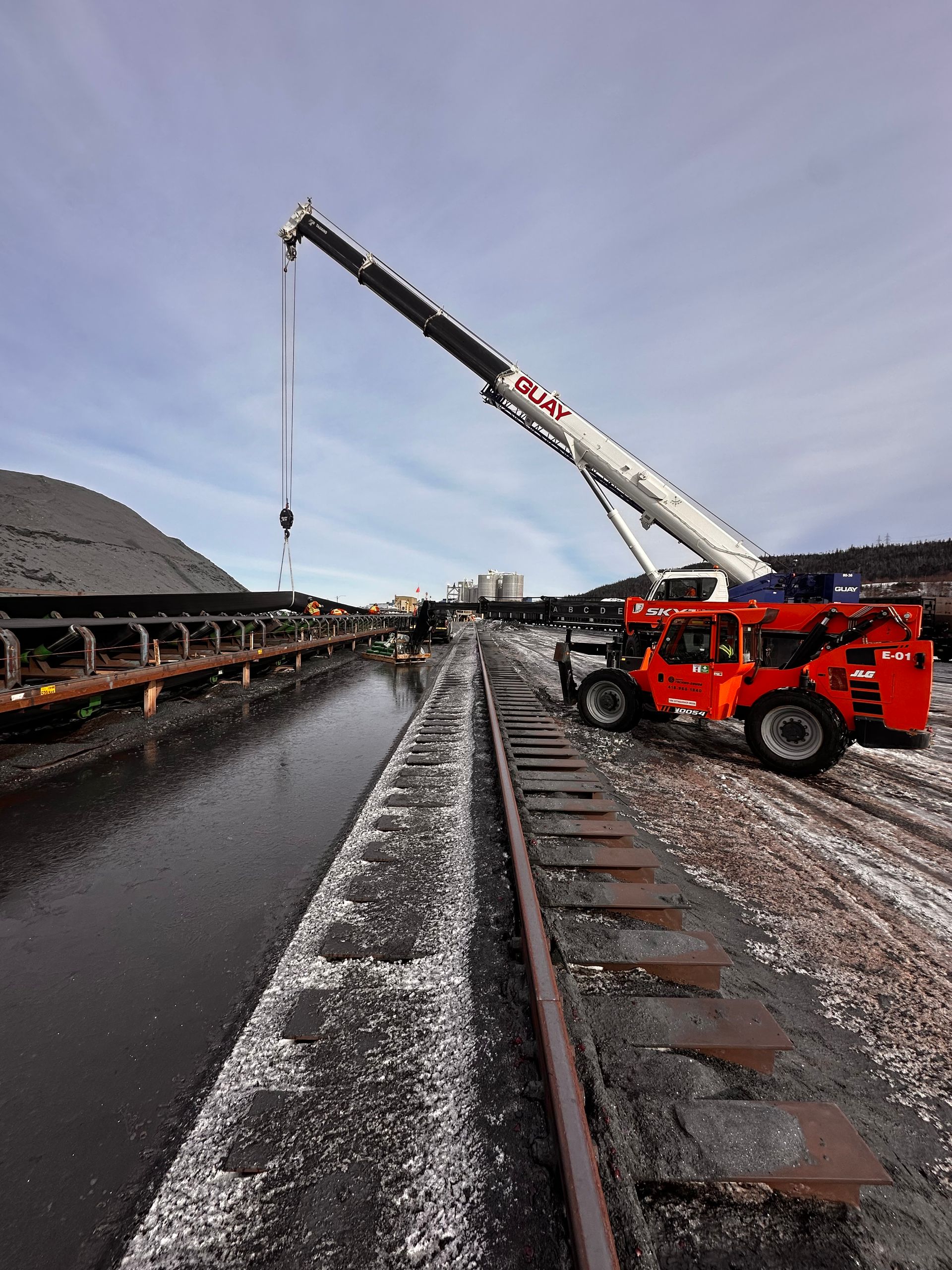 Camion-grue rouge soulevant une charge au bord d'un canal et d'une voie ferrée sous un ciel nuageux