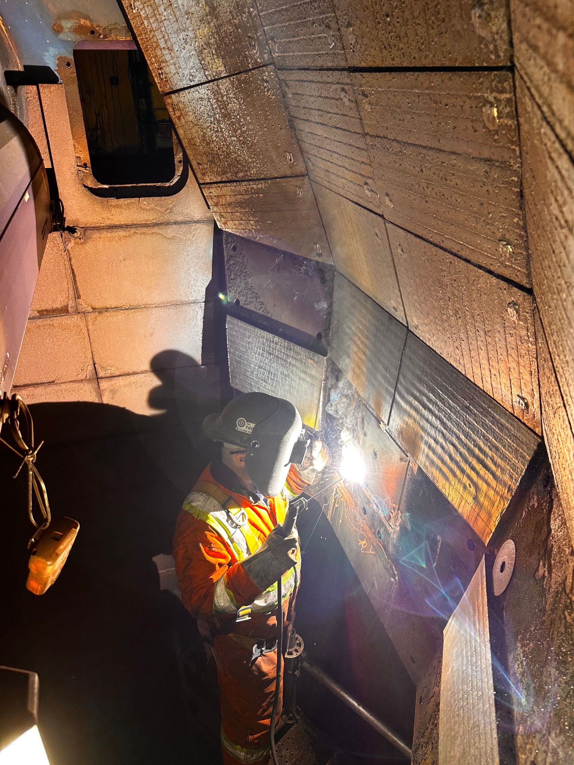 Un ouvrier en tenue de sécurité orange soude à l'intérieur d'un puits industriel étroit et faiblement éclairé.
