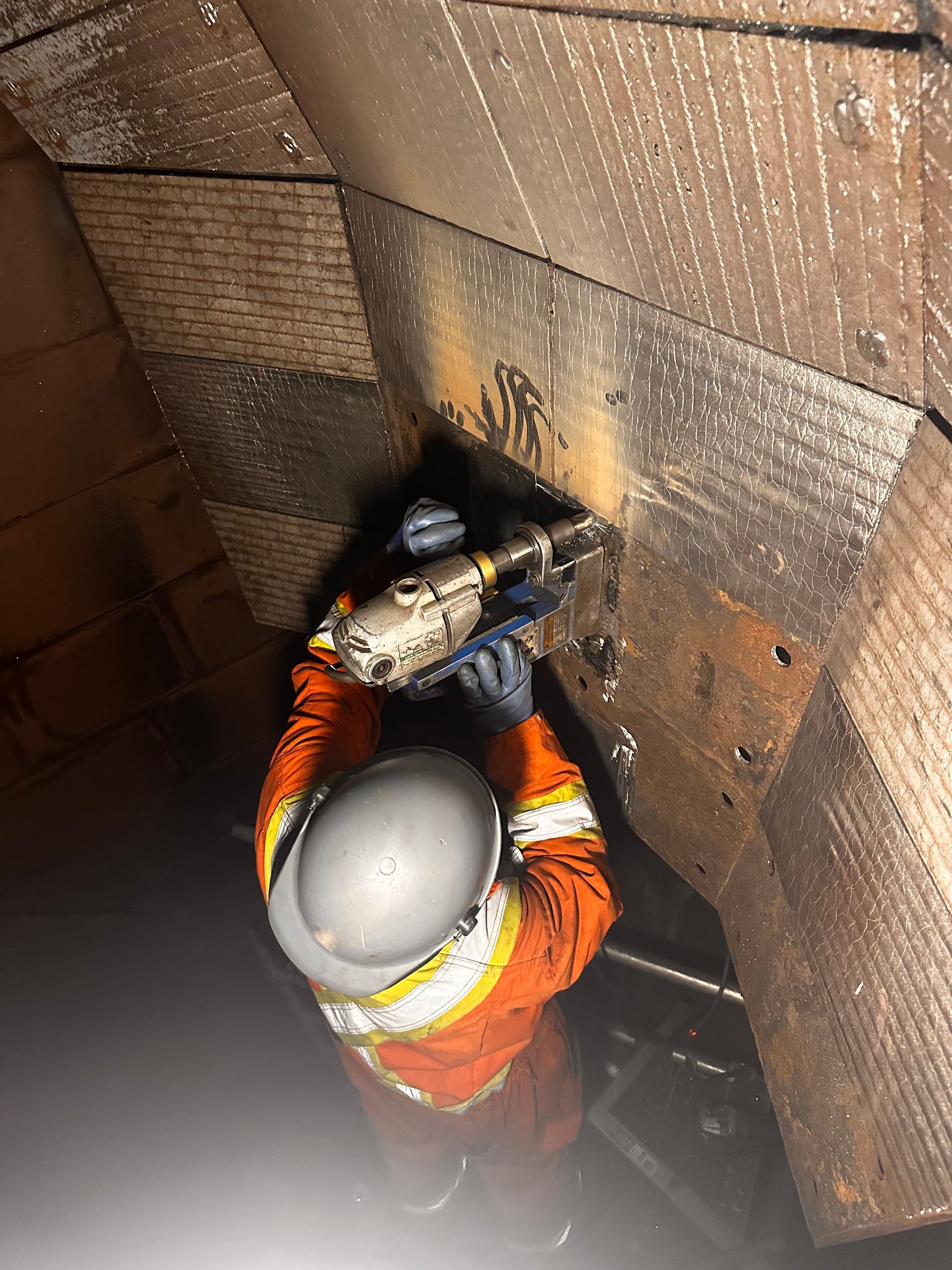 Ouvrier en tenue de sécurité orange utilisant un outil électrique à l'intérieur d'un étroit puits en bois