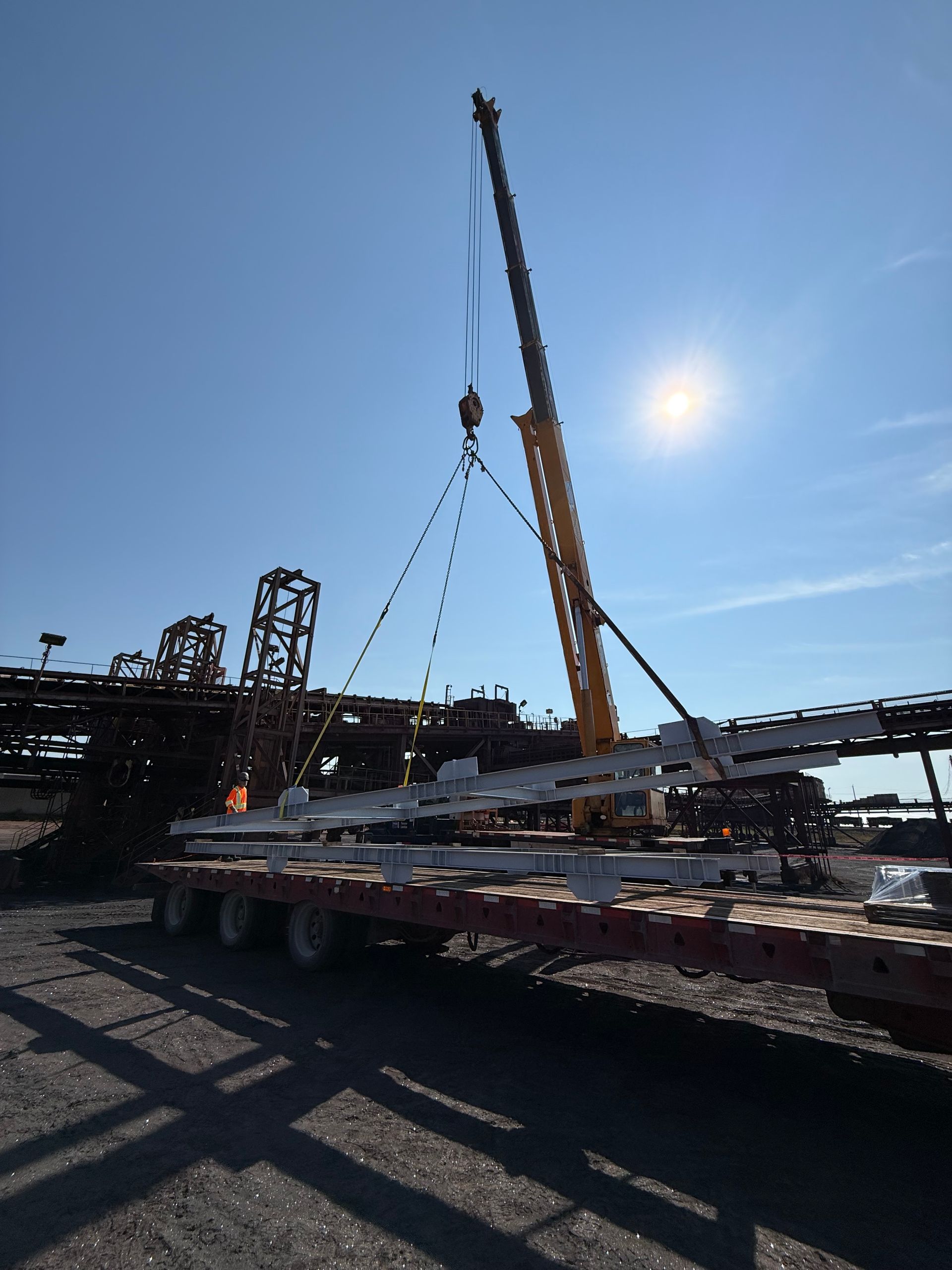 Une grue soulève une grande plateforme industrielle sur un camion plateau sous un ciel d'un bleu éclatant