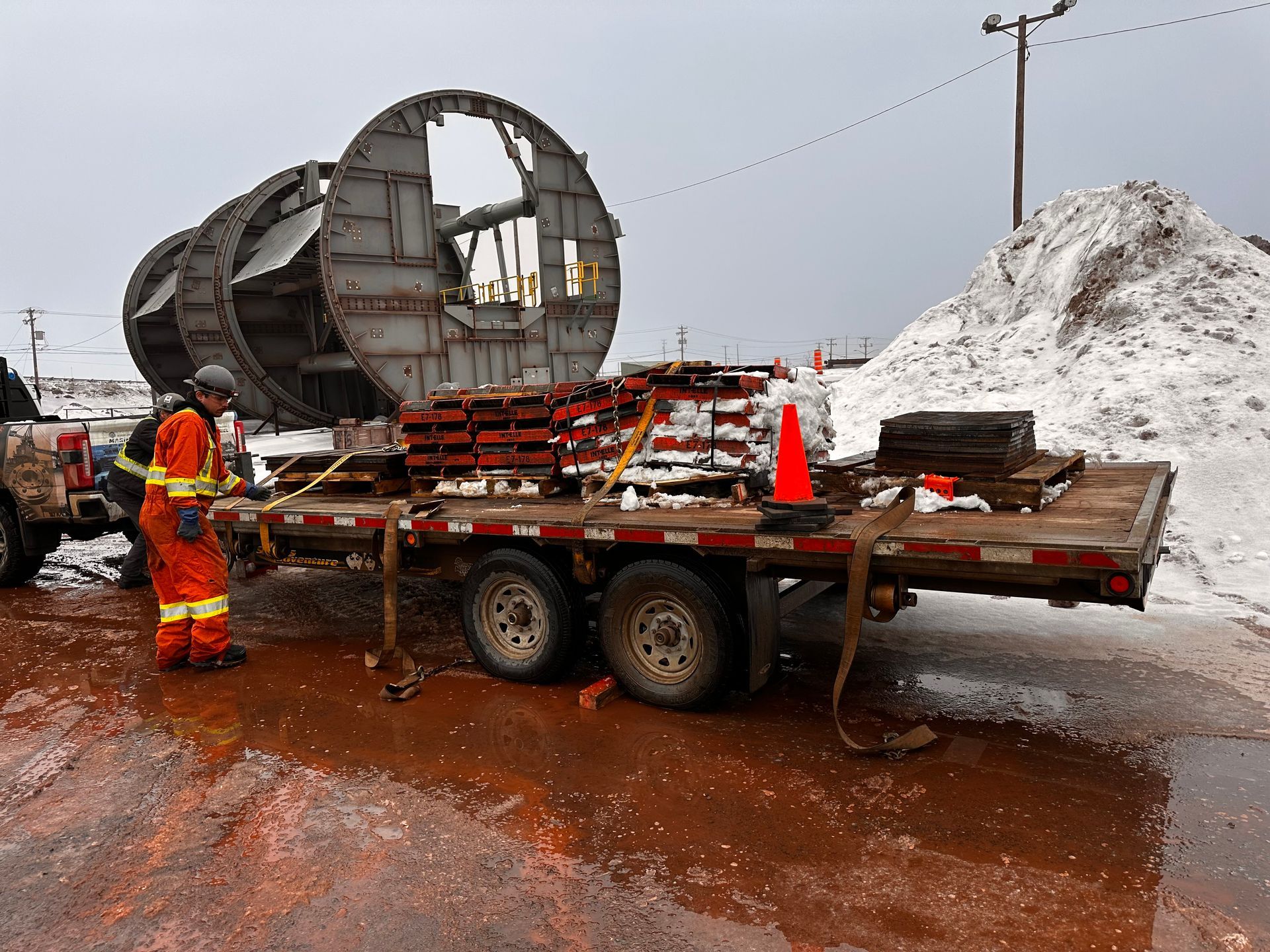 Une équipe de travailleurs routiers se trouve à côté d'un camion plateau transportant un gros cylindre métallique sur un chantier enneigé et boueux.