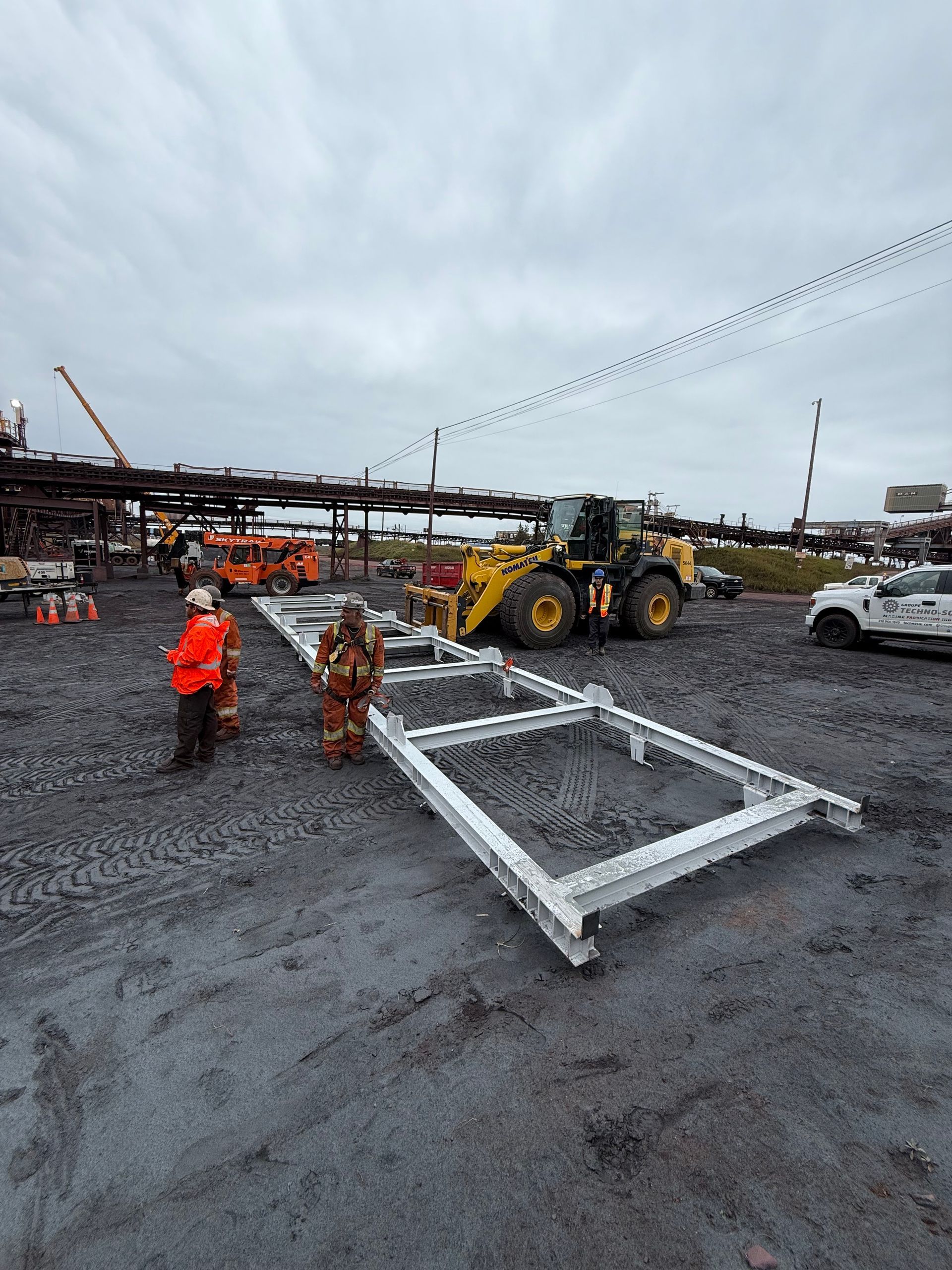 Chantier de construction avec des ouvriers, une pelleteuse et une grande structure métallique sur un sol humide sous un ciel couvert
