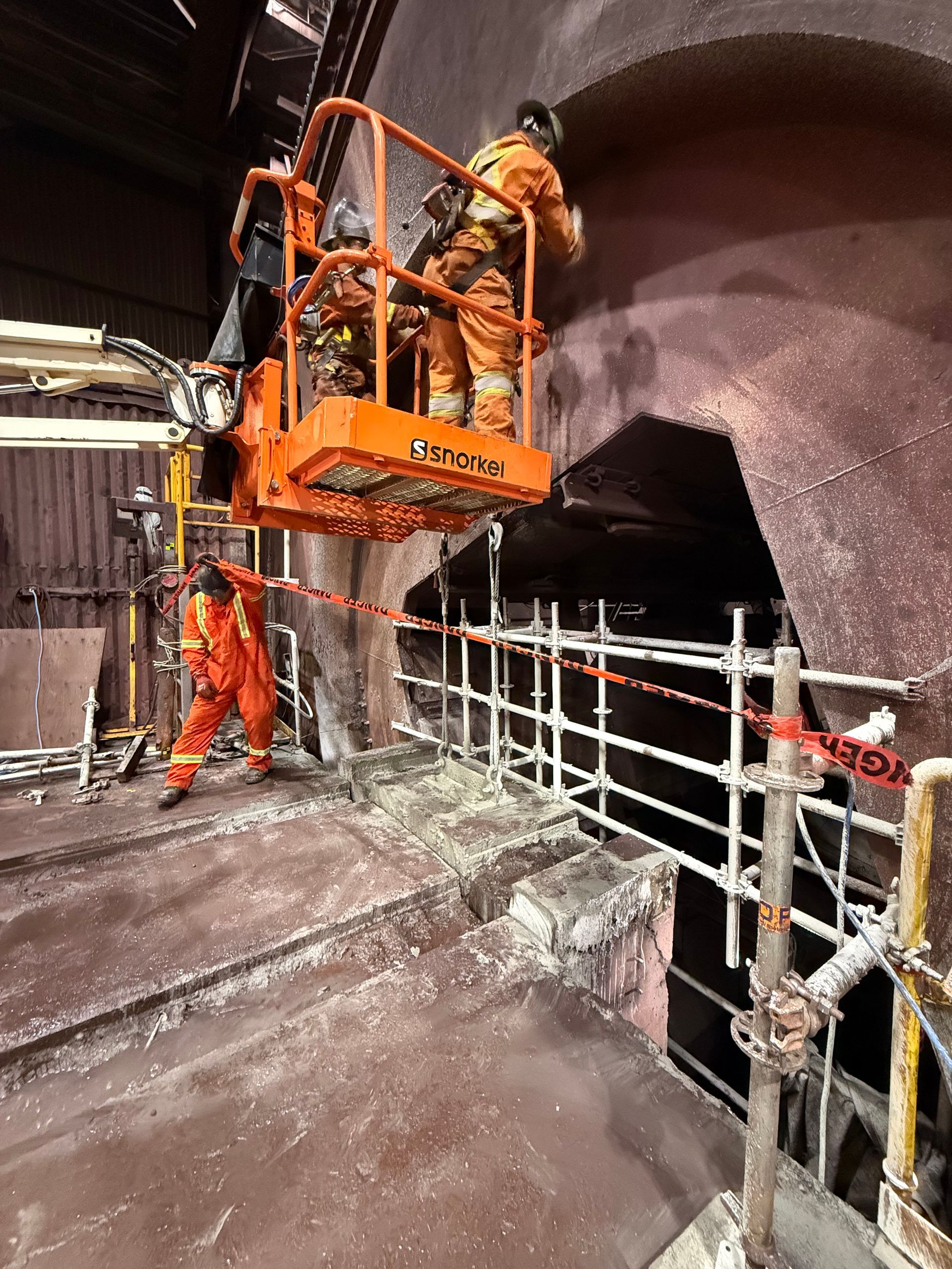 Deux ouvriers en tenue de sécurité orange inspectent, depuis une nacelle, la paroi humide d'un tunnel, à côté des garde-corps.