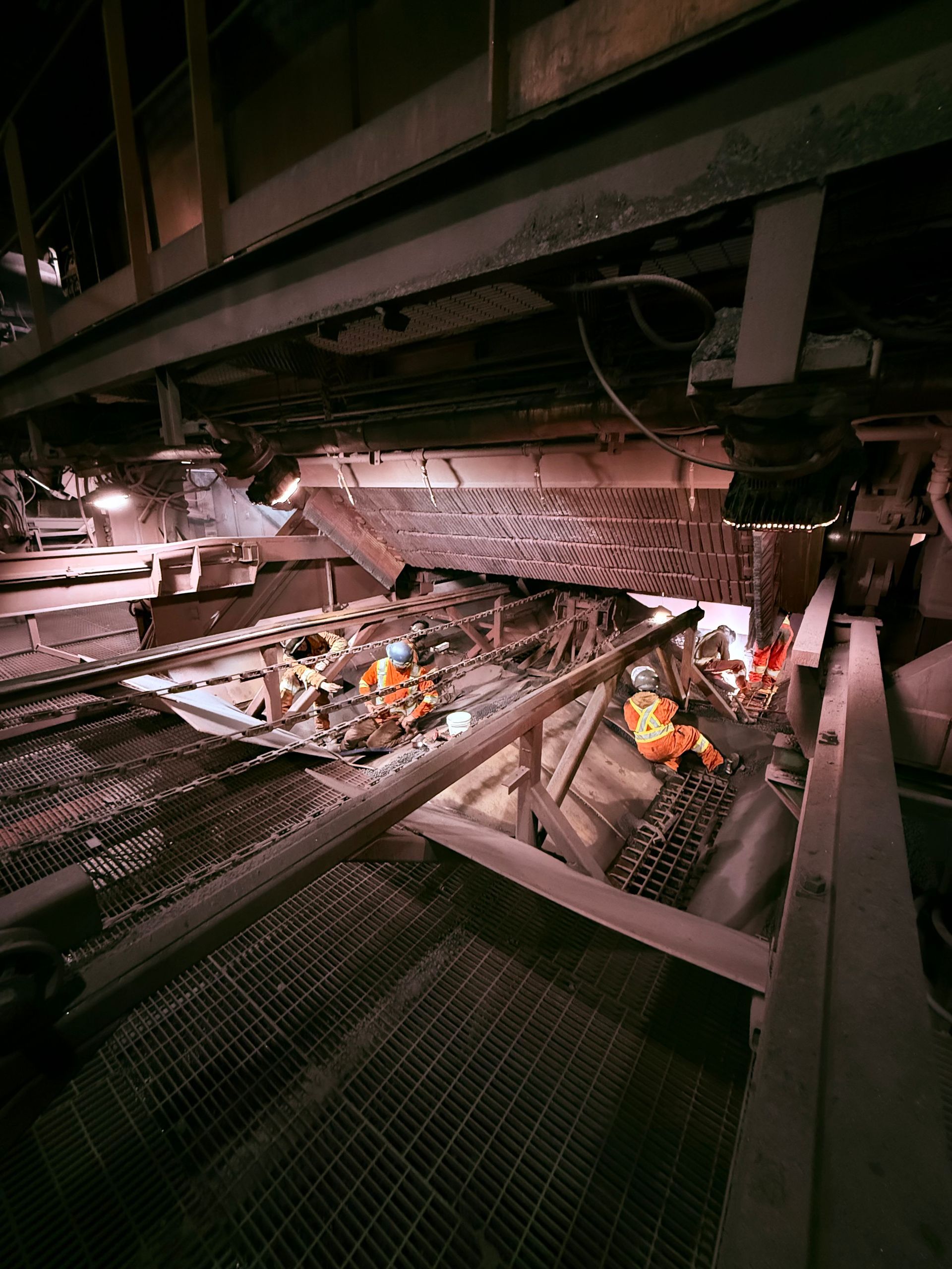 Des passerelles métalliques surplombent une fosse industrielle obscure, avec deux ouvriers en tenue de sécurité orange en dessous.