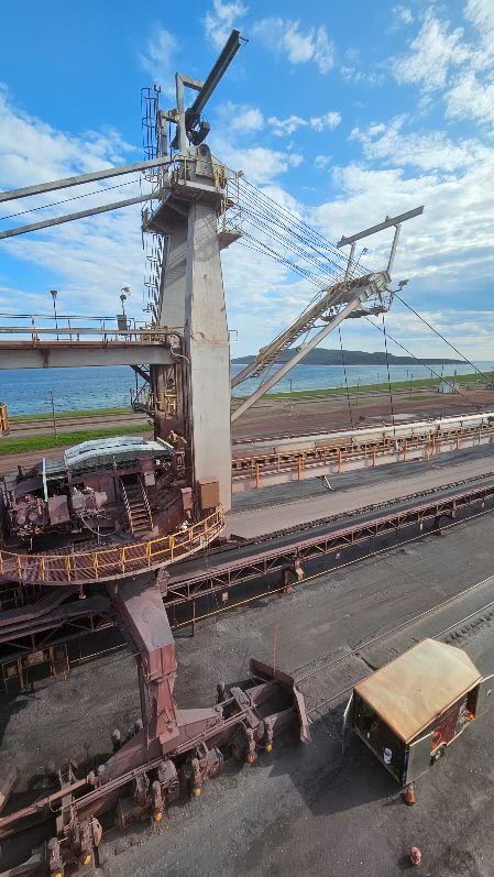 Chantier de construction industrielle avec une haute tour en béton, des poutres en acier et des voies ferrées sous un ciel bleu.