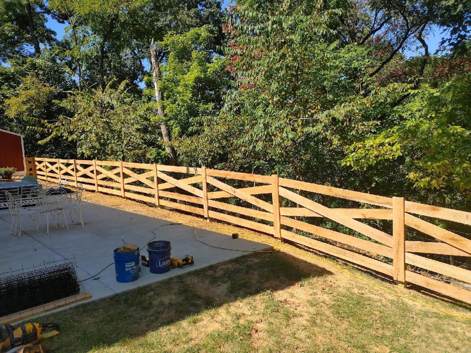 Wooden fence with cross-bracing in a backyard. Blue buckets and tools sit on the patio. Trees in background.