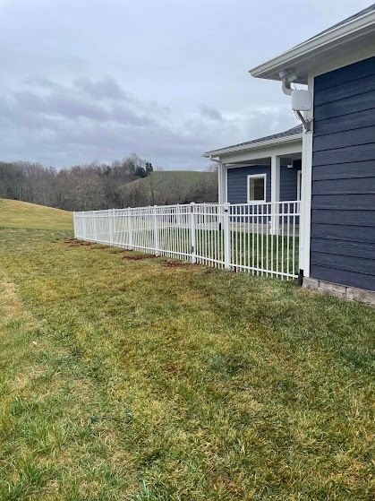 White fence along a house, framing a lawn and rolling hills under an overcast sky.