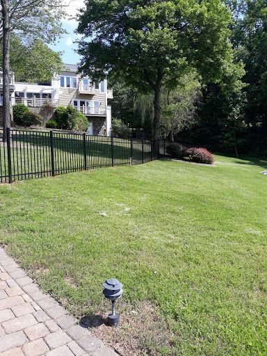 Lawn with black fence, trees, and a house in the background. A brick patio is in the foreground.
