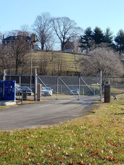Gated driveway with cars behind a chain-link fence, a vending machine on the left, and a grassy hill in the background.