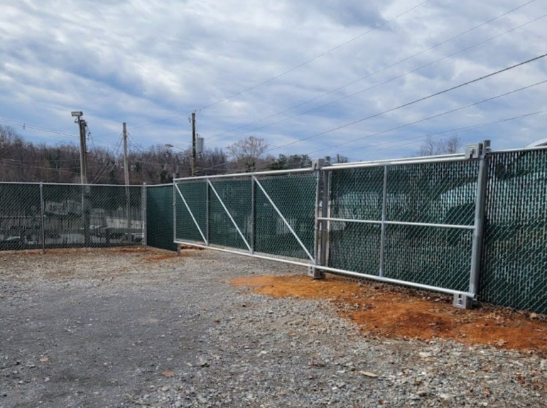 Chain link fence with a gate, covered in green screening, on a gravel lot under a cloudy sky.