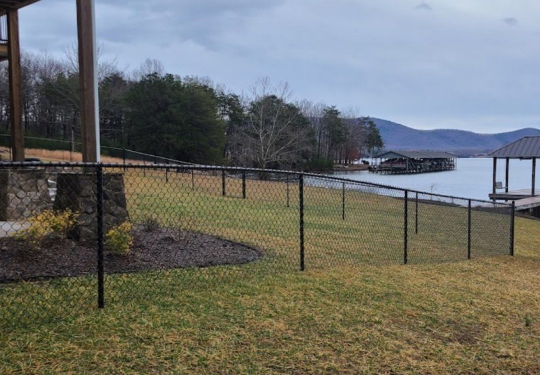 Black chain-link fence on grassy yard near lake with trees, mountains, and overcast sky.