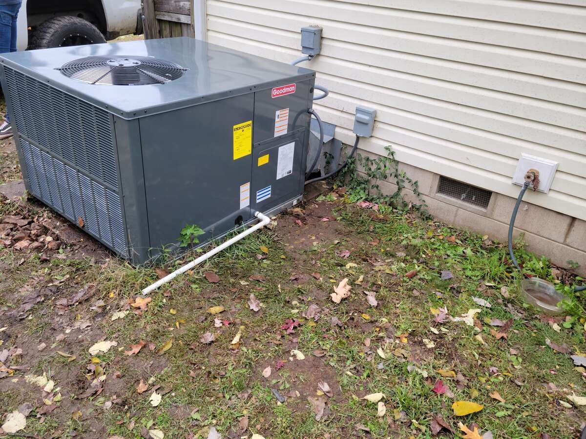 Outdoor air conditioner unit beside a house wall, with hoses and a small utility box nearby.