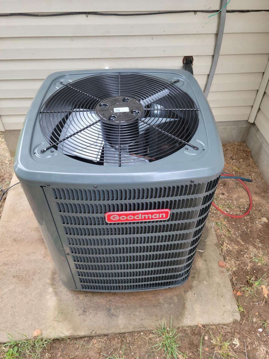 Outdoor air conditioner unit beside a house wall on a concrete pad, with a red hose nearby.