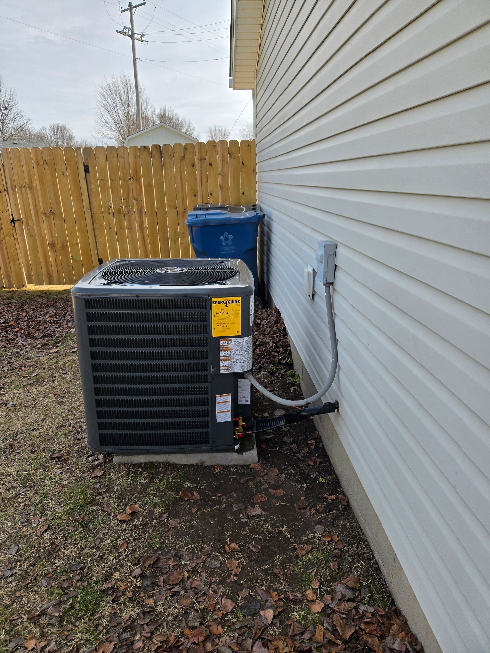Outdoor HVAC unit beside a white house wall and wooden fence, with a blue bin behind it.