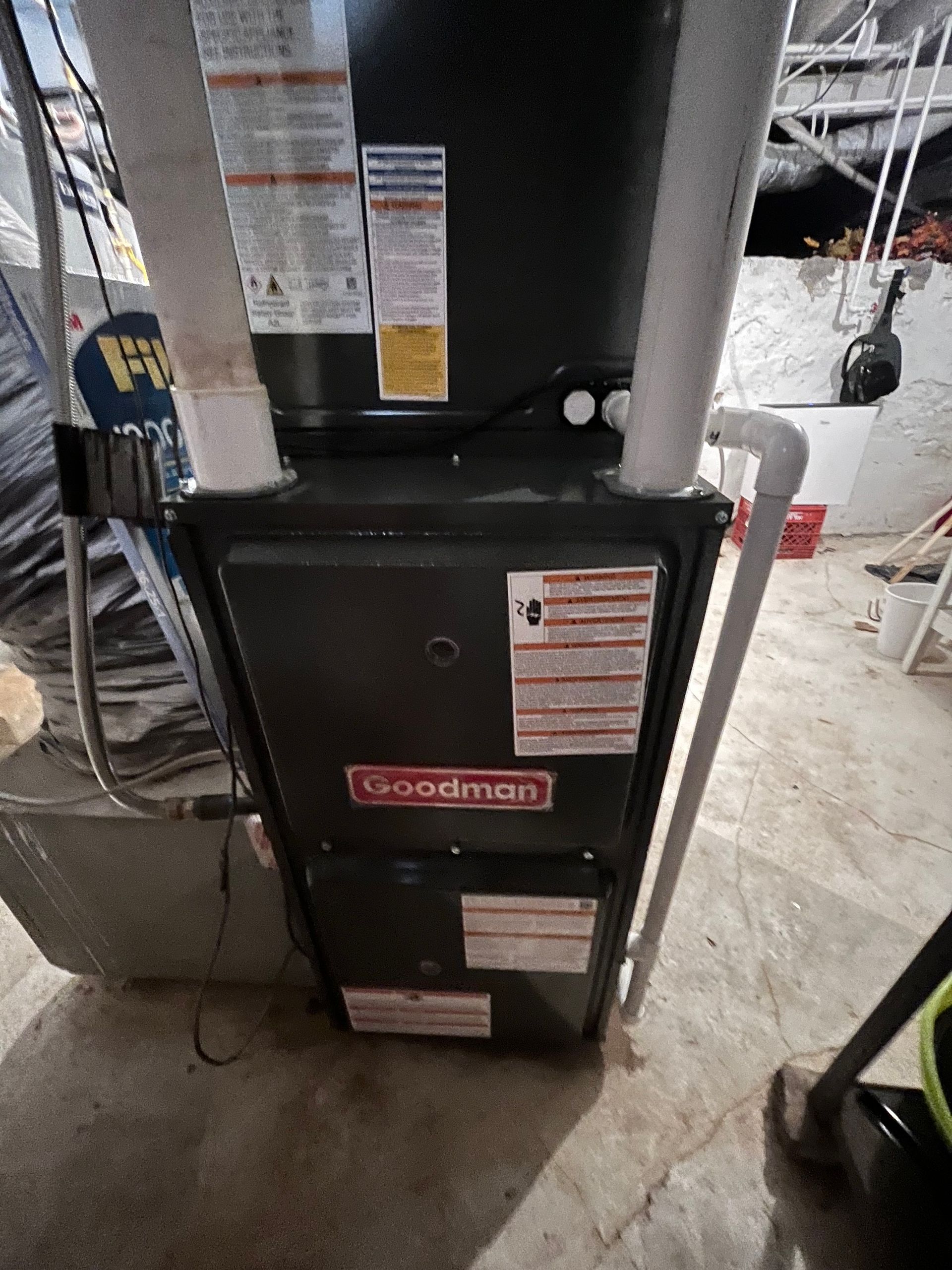 Black Goodman furnace in a basement, with warning labels and a gray vent pipe.