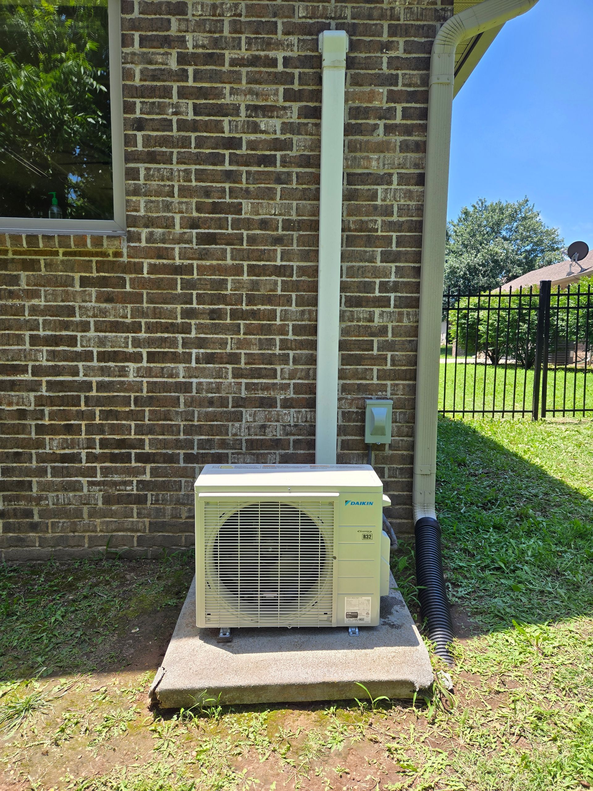 Outdoor HVAC unit beside a brick house wall, with a downspout and a small utility meter nearby
