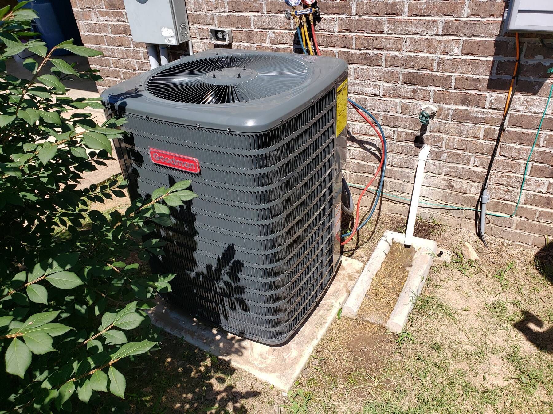 Outdoor air conditioning unit beside a brick house wall, with pipes and hoses in a sunny yard