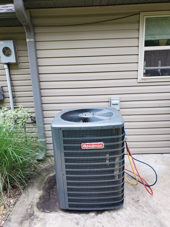 Outdoor central air conditioner beside a beige house, with hoses attached.