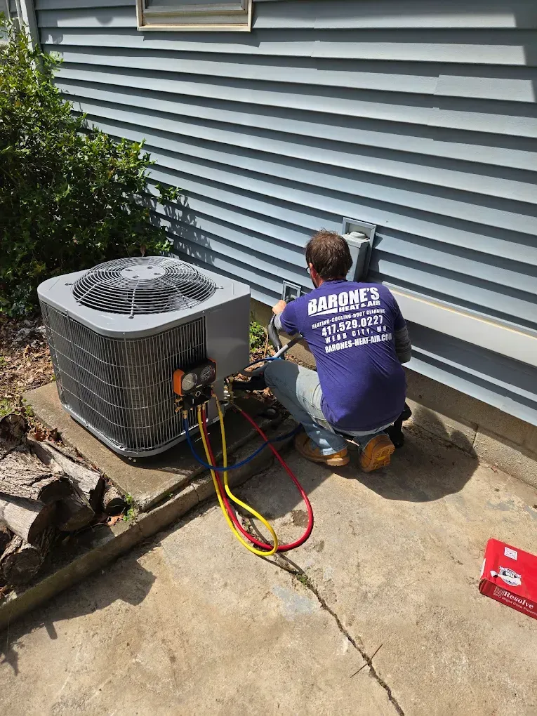 Technician kneels by outdoor AC unit, checking refrigerant gauges beside a house wall.
