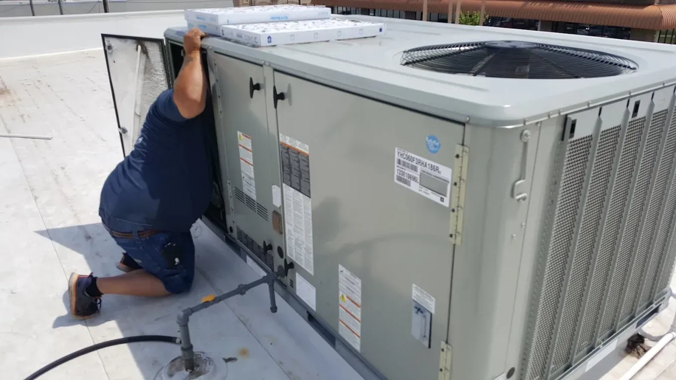 Worker kneeling beside a rooftop HVAC unit, inspecting or servicing it.
