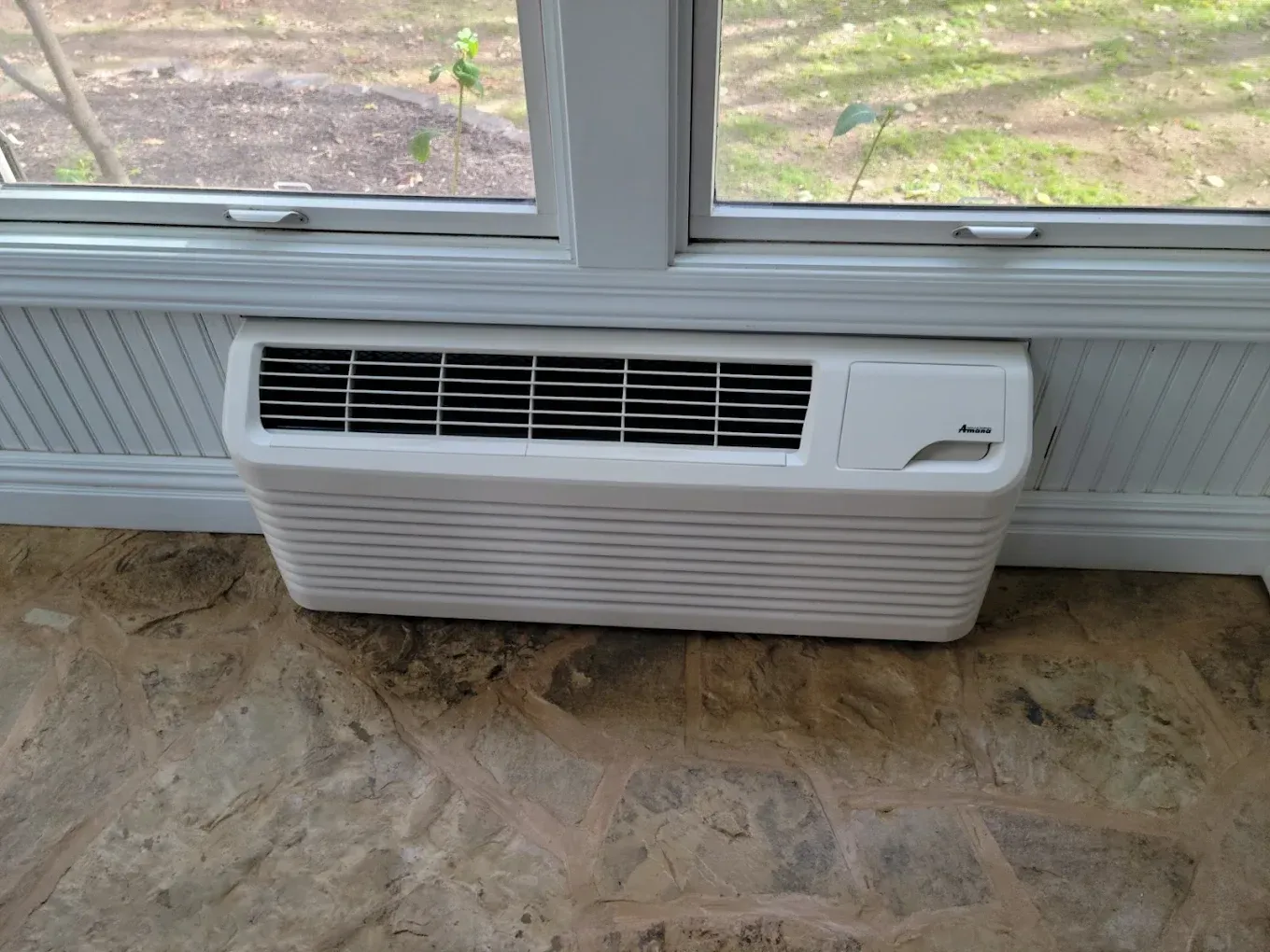 White window air conditioner under a window in a room with a dirty floor.