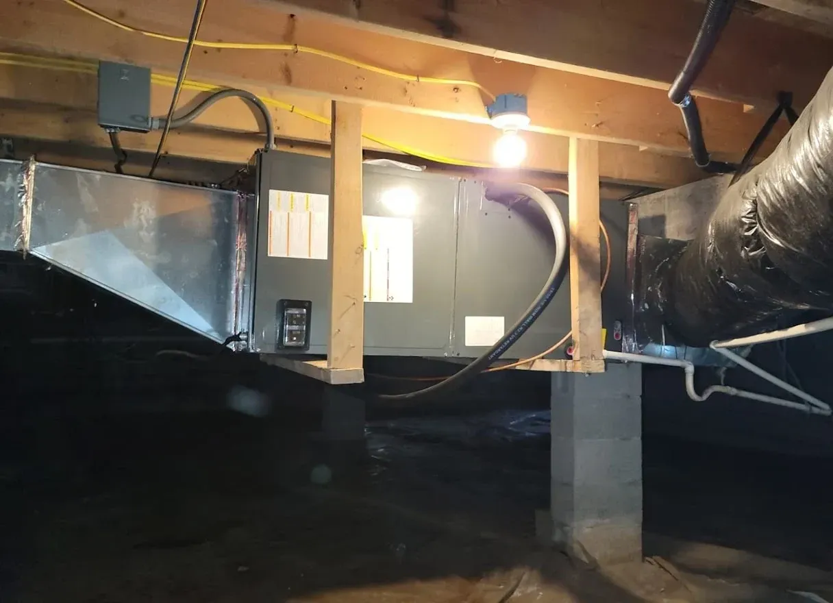 Crawlspace with HVAC ducts, exposed beams, and a furnace unit under a lit floor opening.